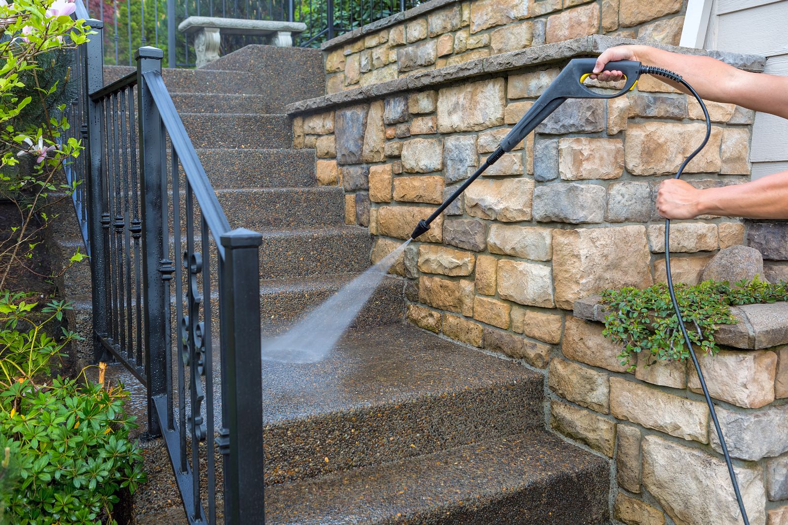 Person pressure washing stone wall and driveway with handheld sprayer