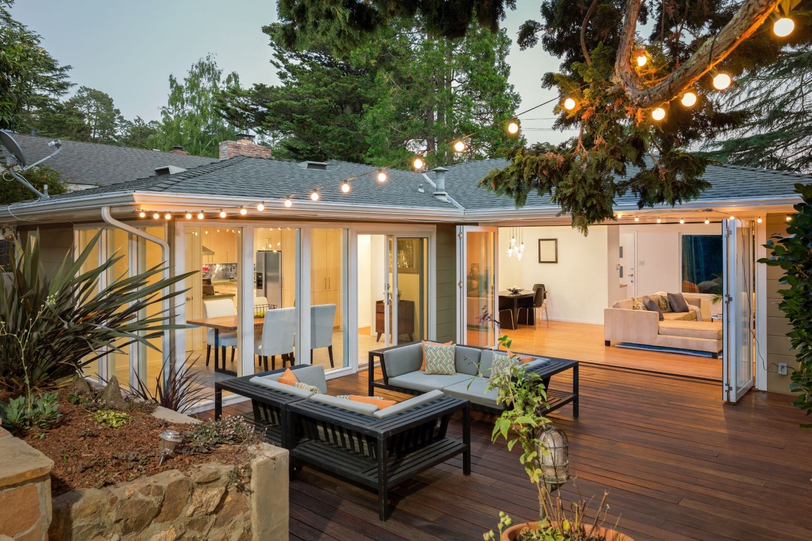 Modern home exterior with illuminated deck, outdoor furniture, and open glass doors at dusk