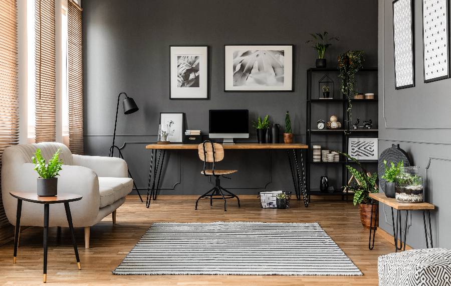 Modern home office with dark gray walls, wooden desk, black chair, white armchair, potted plants, and framed artwork